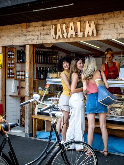 Drei Frauen kaufen an einem Marktstand mit Käse und Feinkost ein, Fahrrad mit Korb im Vordergrund | © Graz Tourismus - Tom Lamm