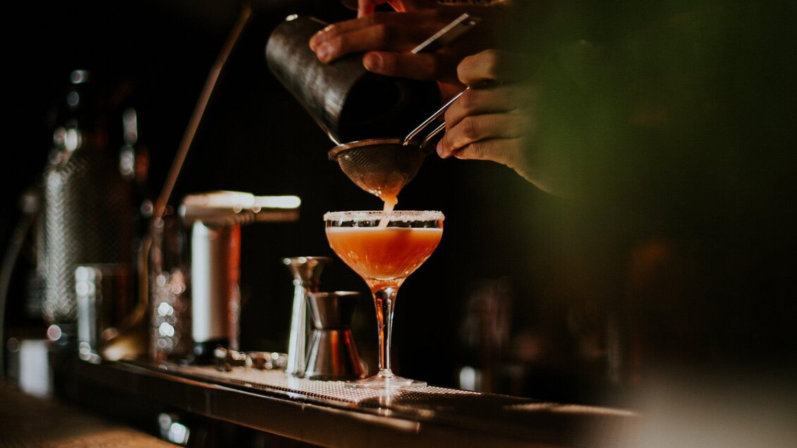 Ein Barkeeper gießt einen Cocktail in ein elegantes Glas. Die Umgebung ist gedämpft und stilvoll beleuchtet. | © Katze Katze - Tinksi