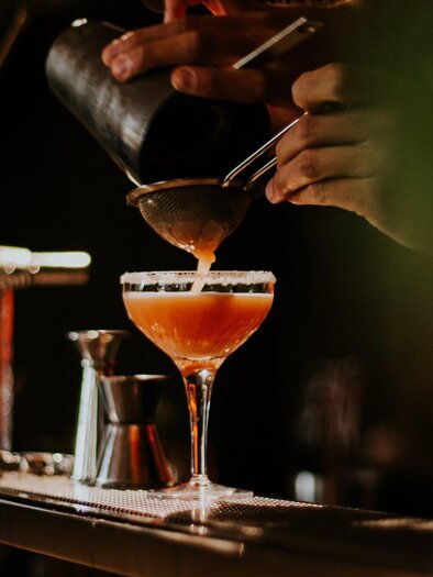 A bartender pours a cocktail into an elegant glass. The surroundings are dim and stylishly lit. | © Katze Katze - Tinksi