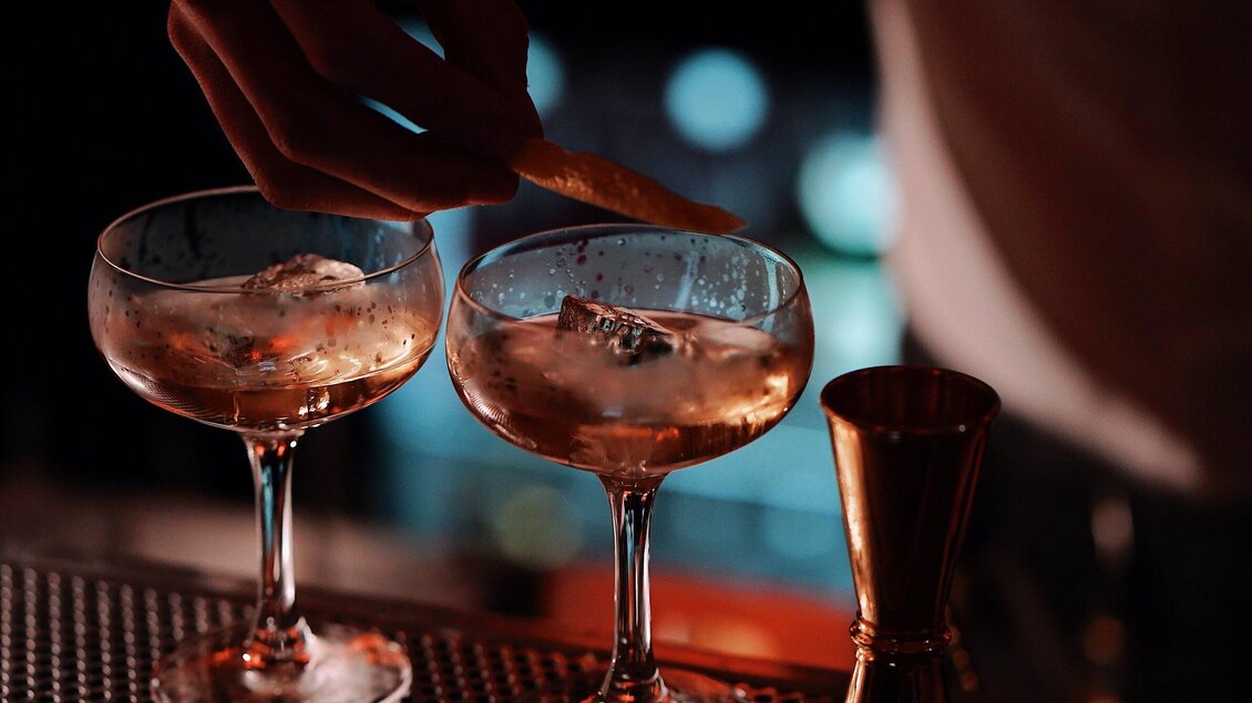 Zwei elegante Cocktails in stilvollen Gläsern stehen auf einer Bartheke. Eine Hand fügt eine Garnitur hinzu und sorgt für einen besonderen Akzent. | © Katze Katze - Leni Tornquist