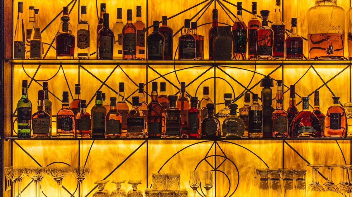 Eine stilvolle Bar mit einer Auswahl an Flaschen in warmem Licht. Im Vordergrund stehen Gläser bereit zum Servieren. | © Katze Katze
