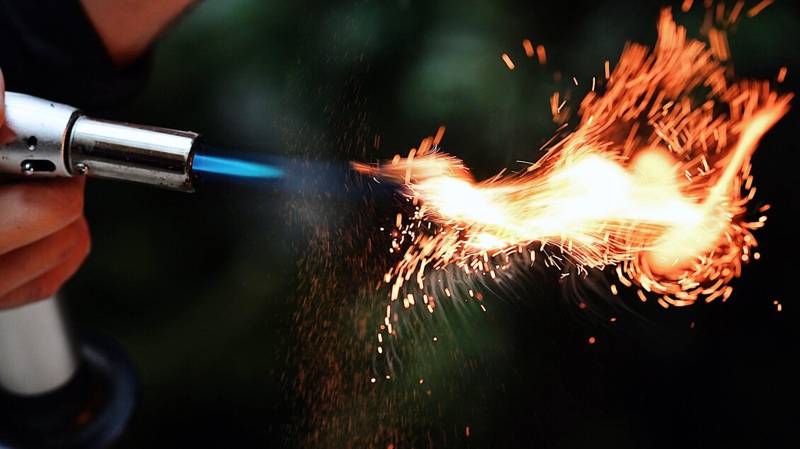 Eine Hand hält einen Brenner, der eine blaue Flamme und Funken erzeugt. Der Hintergrund ist unscharf und grünlich. | © Katze Katze - Leni Tornquist