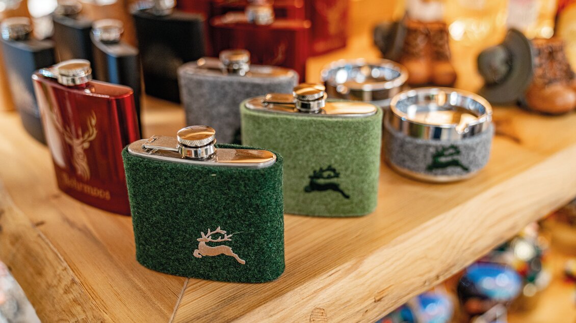 Eine Auswahl an Flachmännern in verschiedenen Farben und Designs auf einem Holzregal. Einige Flachmänner haben tierische Motive und sind aus unterschiedlichem Material gefertigt. | © Christine Höflehner