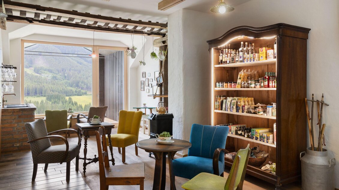 Ein gemütlicher Innenraum mit bunten Stühlen und einem Holzregal, das mit verschiedenen Lebensmitteln gefüllt ist. Große Fenster bieten einen Blick auf die Landschaft. | © Sloho Bergurlaub