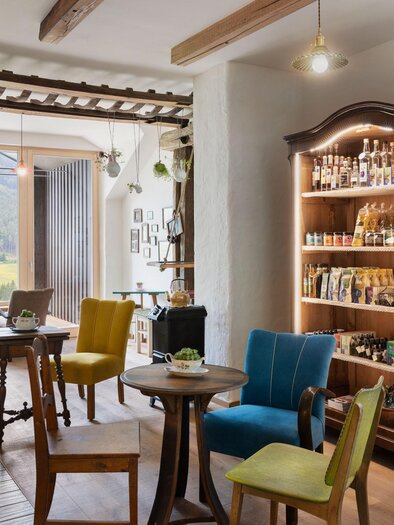 A cozy interior with colorful chairs and a wooden shelf filled with various groceries. Large windows offer a view of the landscape. | © Sloho Bergurlaub
