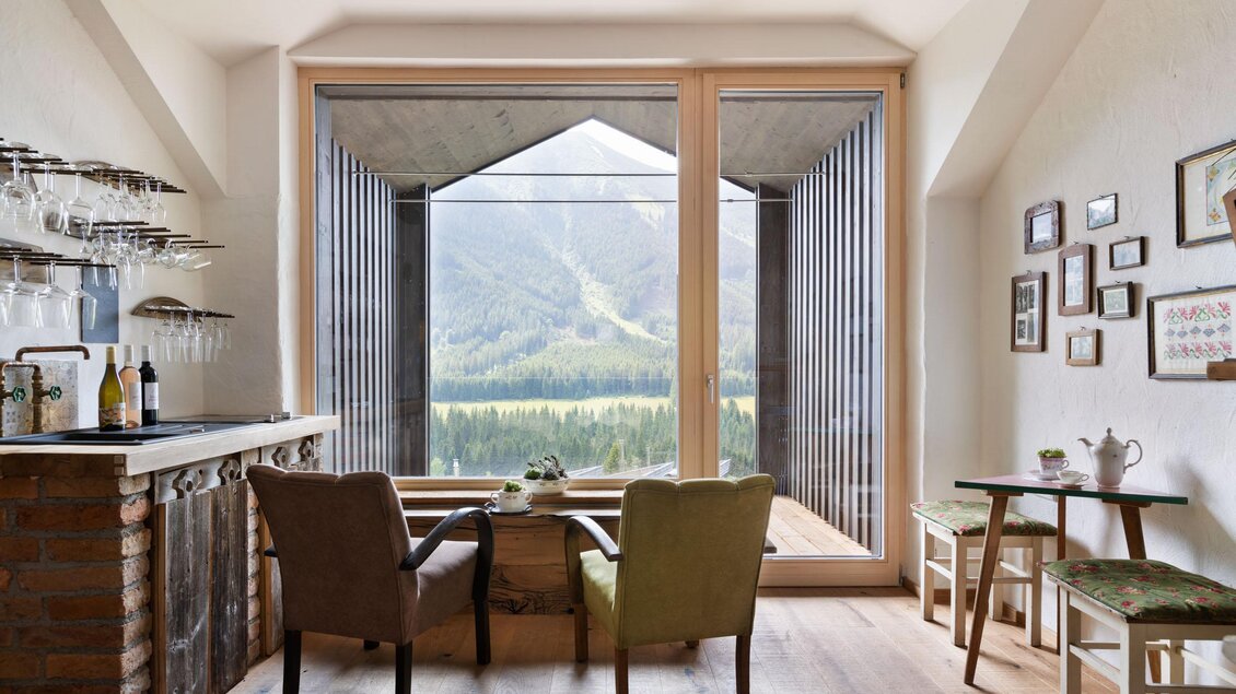 Ein modernes Zimmer mit großen Fenstern, die einen Blick auf die Berge bieten. Es gibt gemütliche Stühle und eine kleine Bar mit Gläsern. | © Sloho Bergurlaub