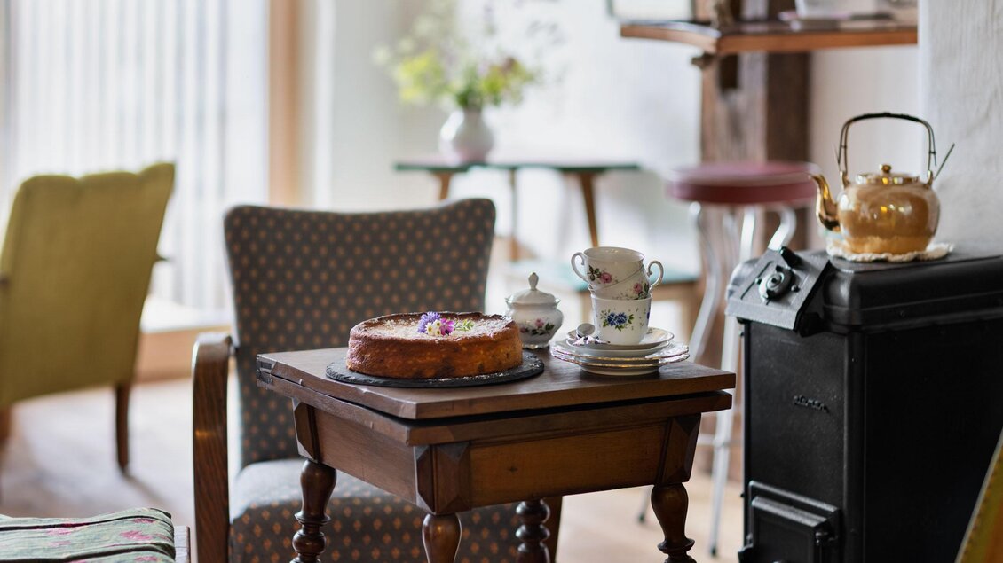 Ein gemütlicher Raum mit einem kleinen Tisch, auf dem ein Kuchen steht. Im Hintergrund sind zwei Sessel und eine Teekanne zu sehen. | © Sloho Bergurlaub