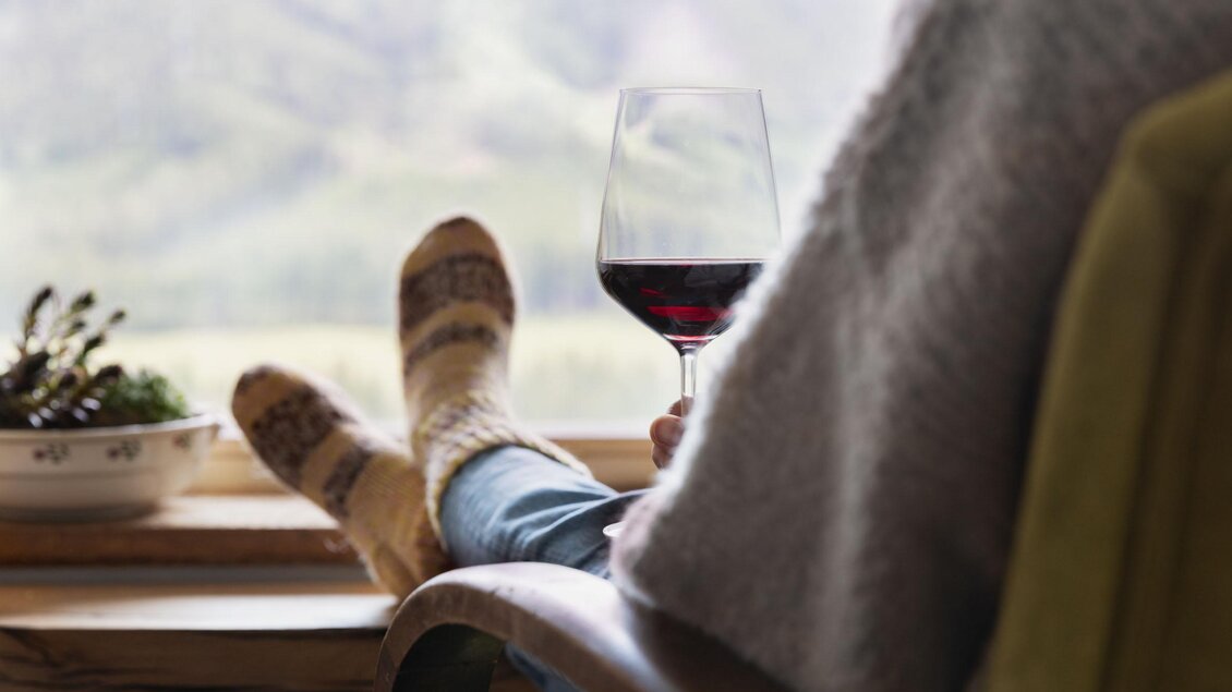 Eine Person sitzt entspannt in einem Sessel und hält ein Glas Rotwein. Im Hintergrund sind grüne Berge und eine ruhige Landschaft zu sehen. | © Sloho Bergurlaub