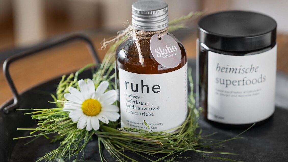Ein Glasfläschchen mit der Aufschrift "ruhe" steht neben einem schwarzen Behälter mit der Beschriftung "heimische superfoods". Umgeben von frischen Kräutern und einer Margerite vermittelt das Bild ein Gefühl von Natürlichkeit und Entspannung. | © Sloho Bergurlaub