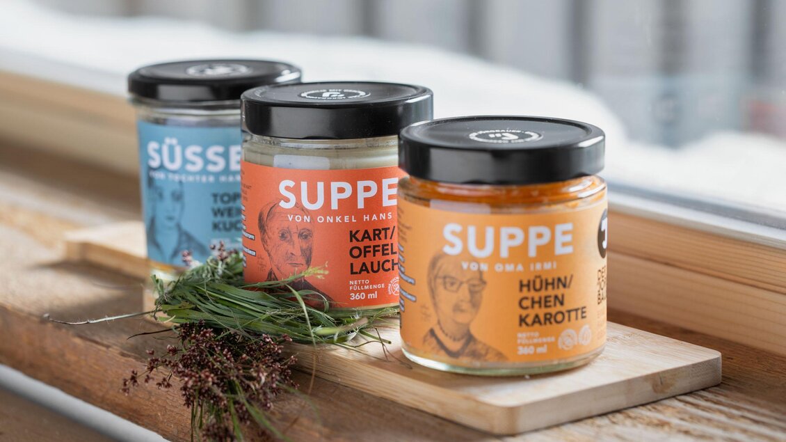 Drei verschiedene Gläser Suppe stehen auf einem Holzbrett. Die Etiketten zeigen Geschmacksrichtungen wie Huhn, Karotte und Süßkartoffel. | © Sloho Bergurlaub