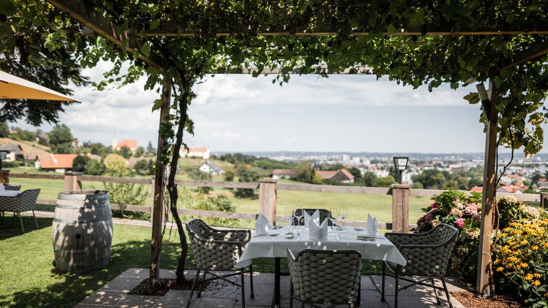 Ein gemütlicher Außenbereich mit einem Tisch und Stühlen unter einem Weinreben-Dach. Im Hintergrund erstreckt sich eine schöne Landschaft mit Blick auf die Stadt. | © Kehlberghof