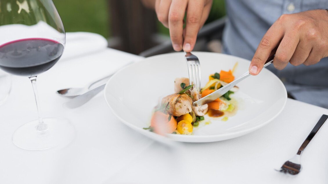 Eine Person schneidet ein schönes Gericht mit Gemüse und Fleisch auf einem Teller. Im Hintergrund steht ein Glas Rotwein. | © Kehlberghof