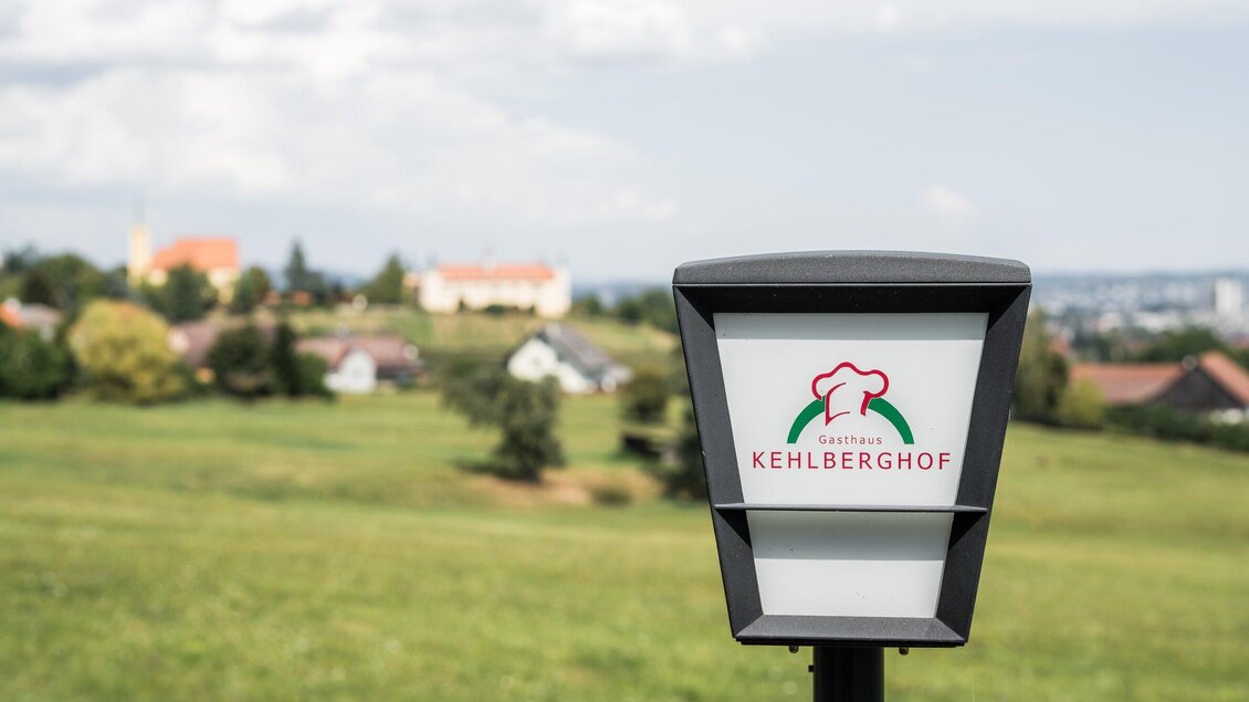 Ein Schild mit dem Logo des Kehberghofs steht vor einer grünen Landschaft. Im Hintergrund sind kleine Häuser und ein blauer Himmel sichtbar. | © Kehlberghof