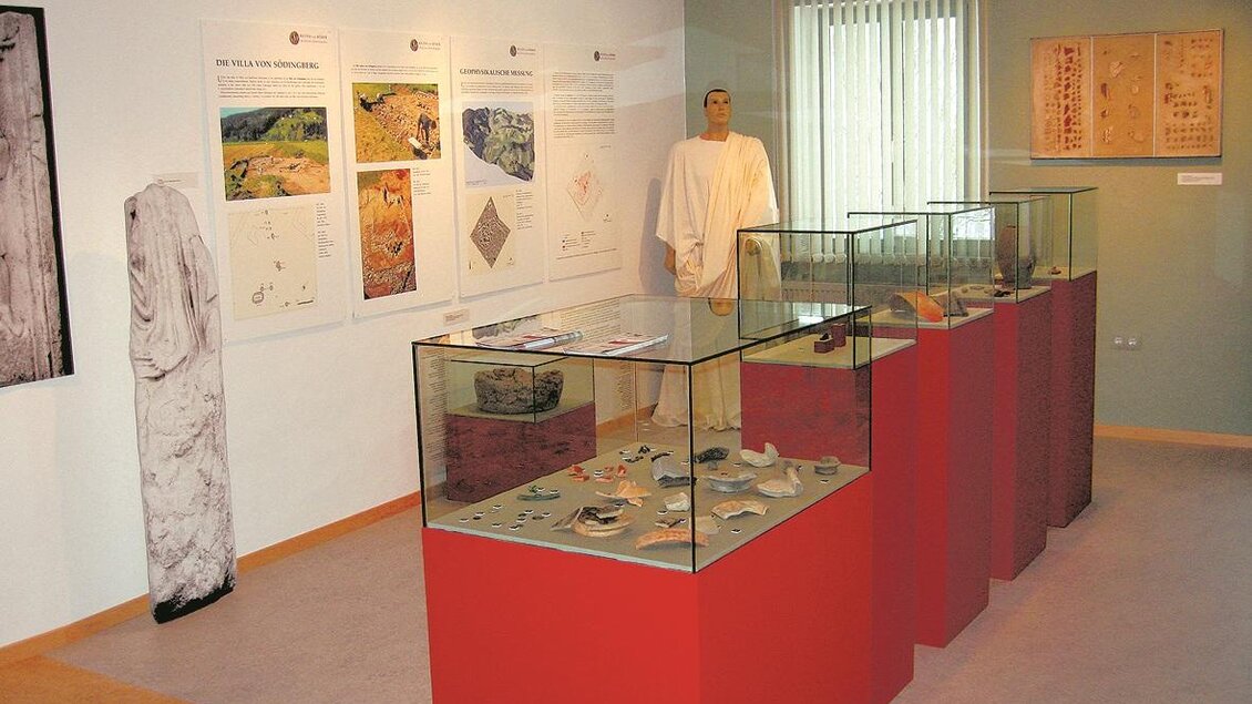 Ein Ausstellungsraum mit Vitrinen, die archäologische Funde zeigen. An der Wand hängen informative Plakate und ein Ausstellungsstück steht in einem Raum mit Tageslicht. | © Gemeinde Geistthal-Södingberg