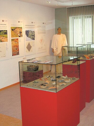 Ein Ausstellungsraum mit Vitrinen, die archäologische Funde zeigen. An der Wand hängen informative Plakate und ein Ausstellungsstück steht in einem Raum mit Tageslicht. | © Gemeinde Geistthal-Södingberg