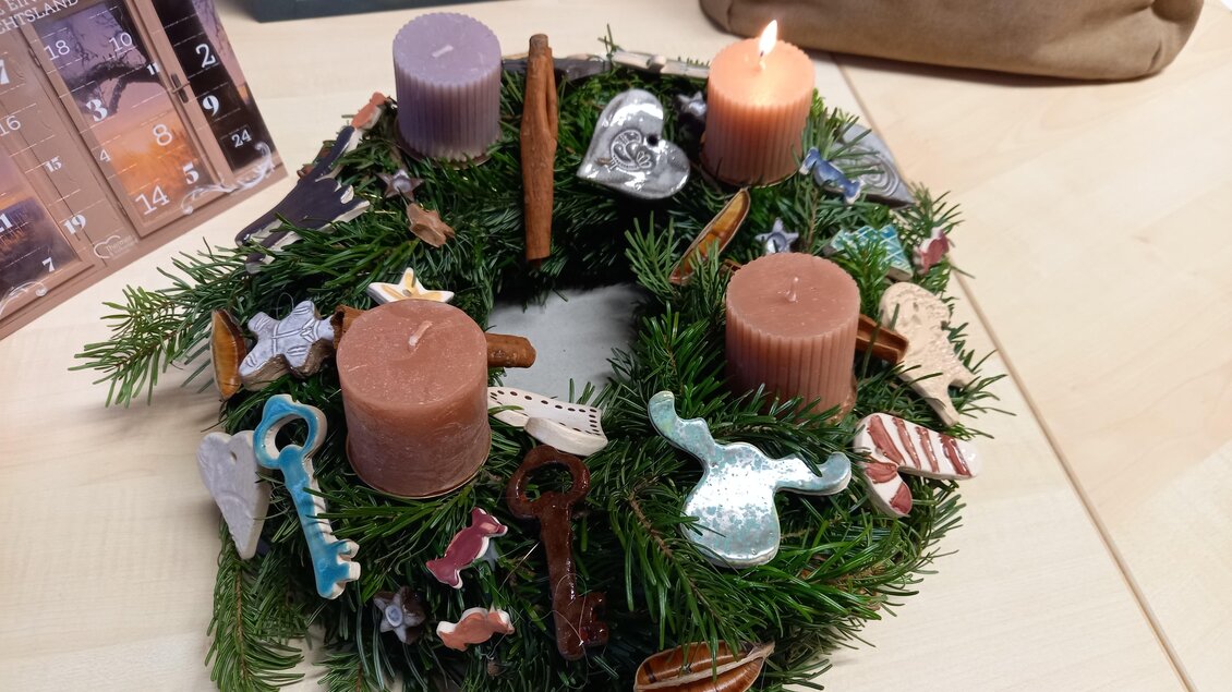 Ein Adventskranz aus Tannenzweigen mit vier Kerzen in verschiedenen Farben. Der Kranz ist mit bunten dekorativen Anhängern geschmückt.