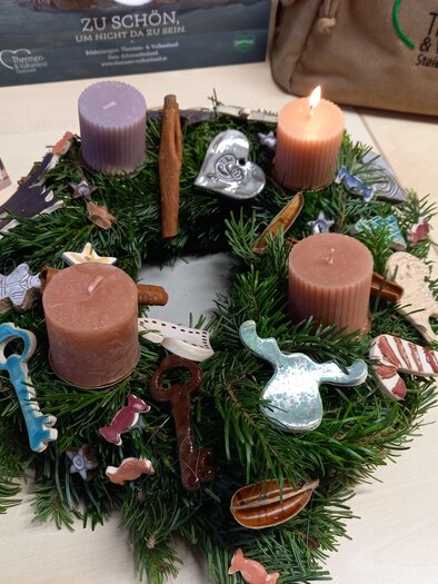 An Advent wreath made of fir branches with four candles in different colors. The wreath is decorated with colorful decorative hangers.