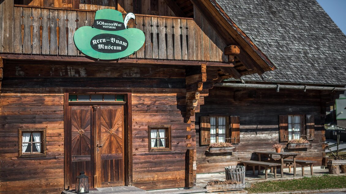 Ein traditionelles, holzverkleidetes Gebäude mit einer grünen Beschilderung. Vor dem Eingang stehen Tische und Stühle in einem ländlichen Umfeld. | © Lipizzanerheimat - Die Abbilderei