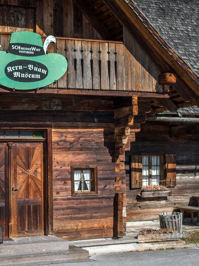 Ein traditionelles, holzverkleidetes Gebäude mit einer grünen Beschilderung. Vor dem Eingang stehen Tische und Stühle in einem ländlichen Umfeld. | © Lipizzanerheimat - Die Abbilderei