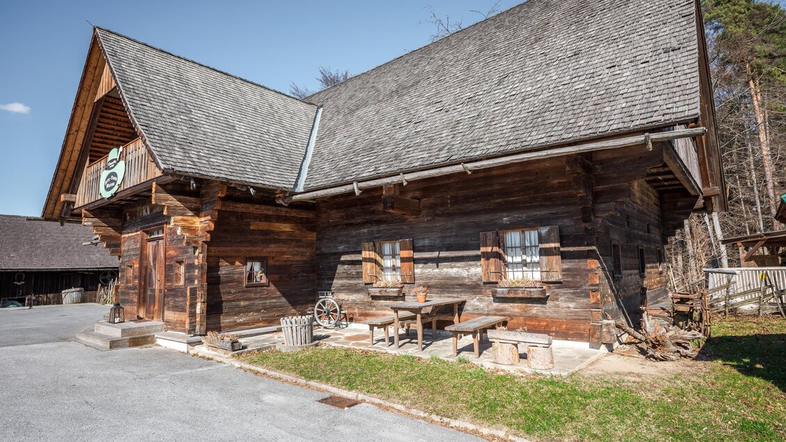 Ein traditionelles Holzhaus mit einem markanten Dach. Vor dem Haus stehen ein Tisch und Stühle auf einem gepflasterten Weg. | © Lipizzanerheimat - Die Abbilderei