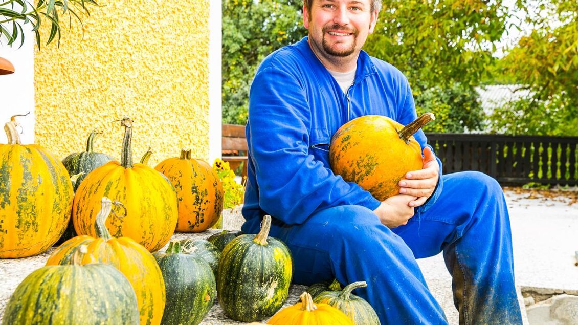 Ein Mann sitzt auf dem Boden und hält einen großen Kürbis. Um ihn herum sind viele Kürbisse in verschiedenen Größen und Farben angeordnet. | © Kernöl Niggas Martin