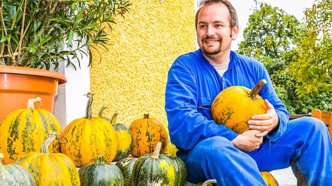 Ein Mann sitzt lächelnd neben einer Sammlung von Kürbissen. Der Hintergrund zeigt eine gelbe Wand und einige Pflanzen. | © Kernöl Niggas Martin