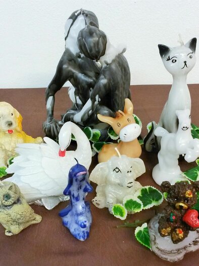 A collection of decorative candles in the shape of animals, including dogs, cats, and a swan. The candles are colorful and stand on a brown background with green leaves. | © Birgit Stockenreitzner
