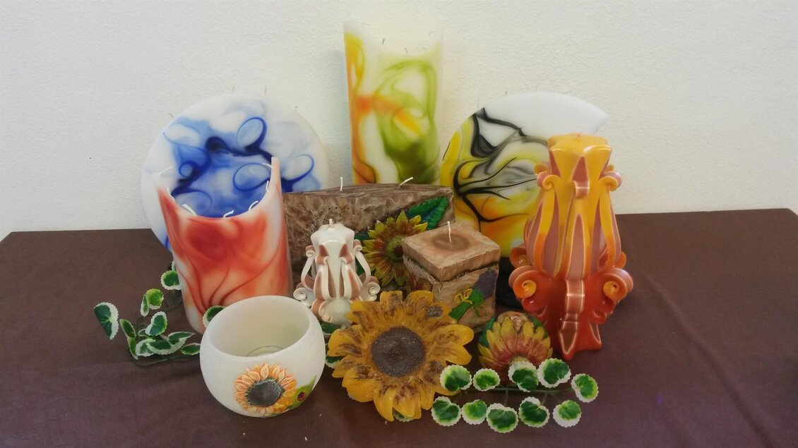 Eine Sammlung von bunten Kerzen in verschiedenen Formen und Farben steht auf einem Tisch. Im Vordergrund sind dekorative Elemente wie Sonnenblumen und grünes Blattwerk zu sehen. | © Birgit Stockenreitner