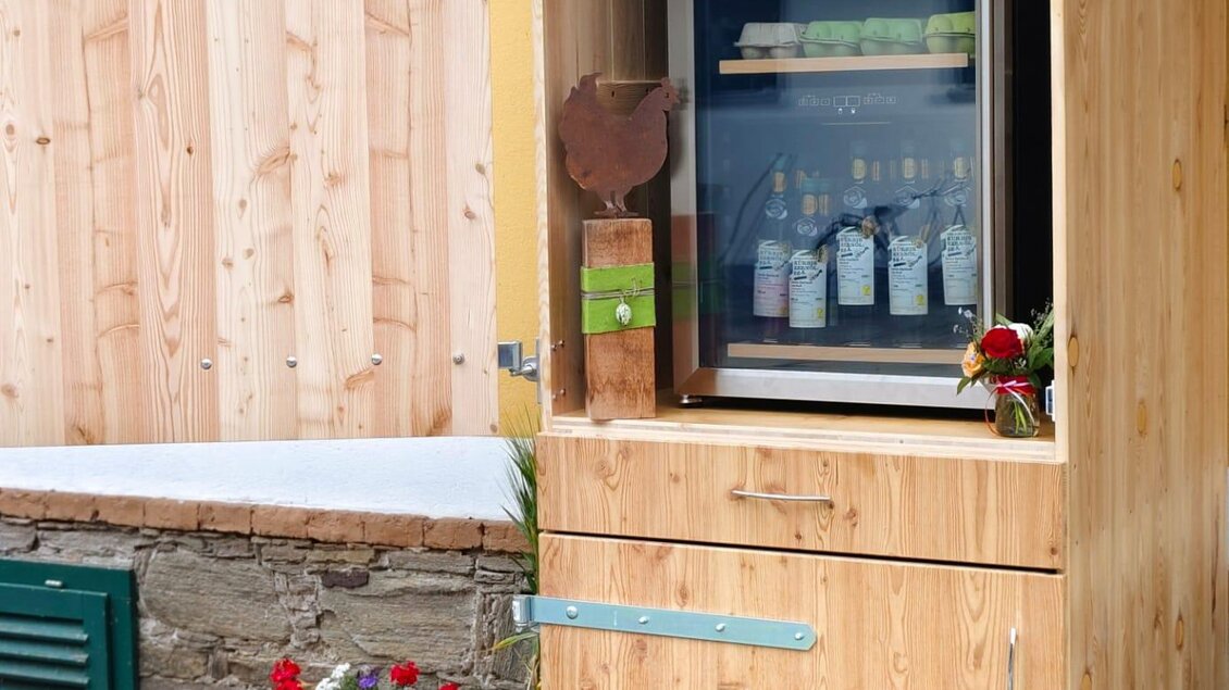 Ein Holzregal mit Kühlschrank steht an einer Hauswand. Darin sind verschiedene Getränke sichtbar, umgeben von bunten Blumen. | © Eberhardt