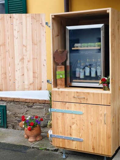 A wooden shelf with a refrigerator stands against a house wall. Various drinks are visible inside, surrounded by colorful flowers. | © Eberhardt