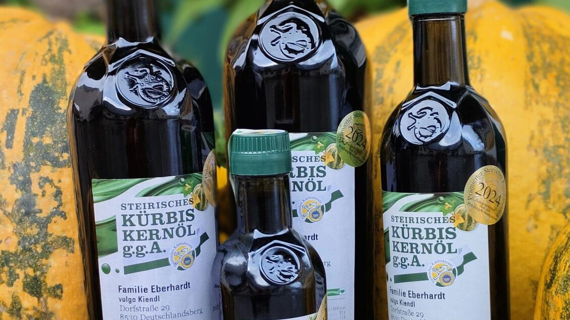 Eine Auswahl an Flaschen mit Kürbiskernöl steht vor großen Kürbissen. Die Flaschen sind in verschiedenen Größen und haben ein ansprechendes Design. | © Eberhardt