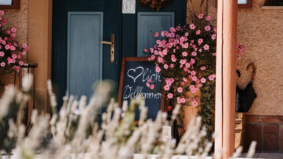 Eine einladende Haustür mit einer Tafel, auf der „Willkommen“ steht. Bunte Blumen stehen in Töpfen neben der Tür. | © Jaglhof