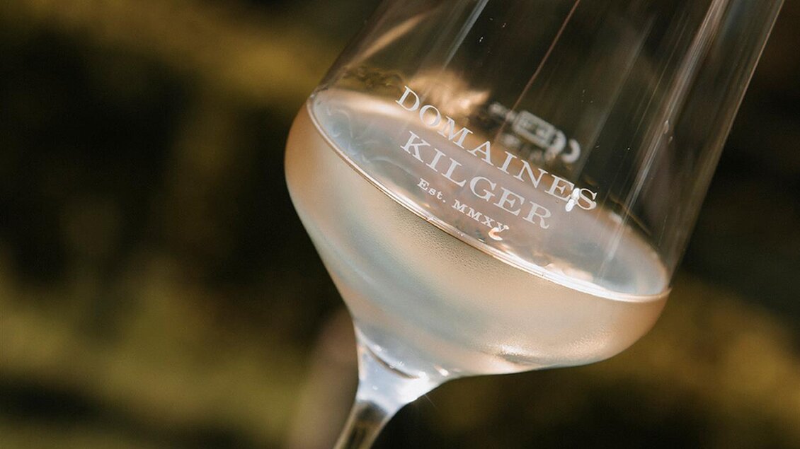 Ein Glas mit hellem Wein steht auf einem Tisch. Im Hintergrund sind grüne Weinberge zu sehen. | © Jaglhof