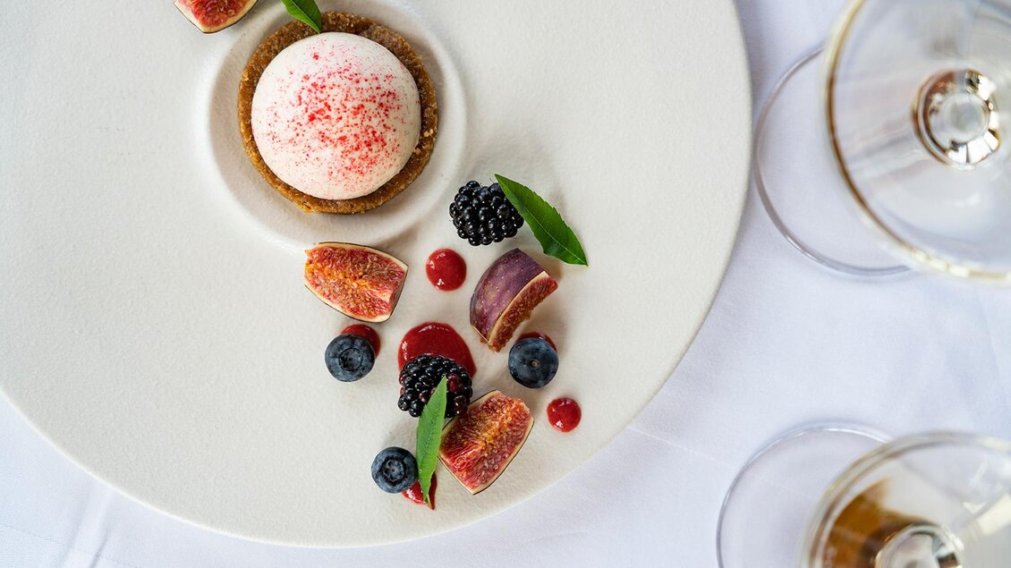 Ein elegantes Dessert auf einem weißen Teller, bestehend aus einer cremigen Mousse und verschiedenen Früchten. Umgeben von Heidelbeeren, Brombeeren und einer fruchtigen Sauce. | © Jaglhof
