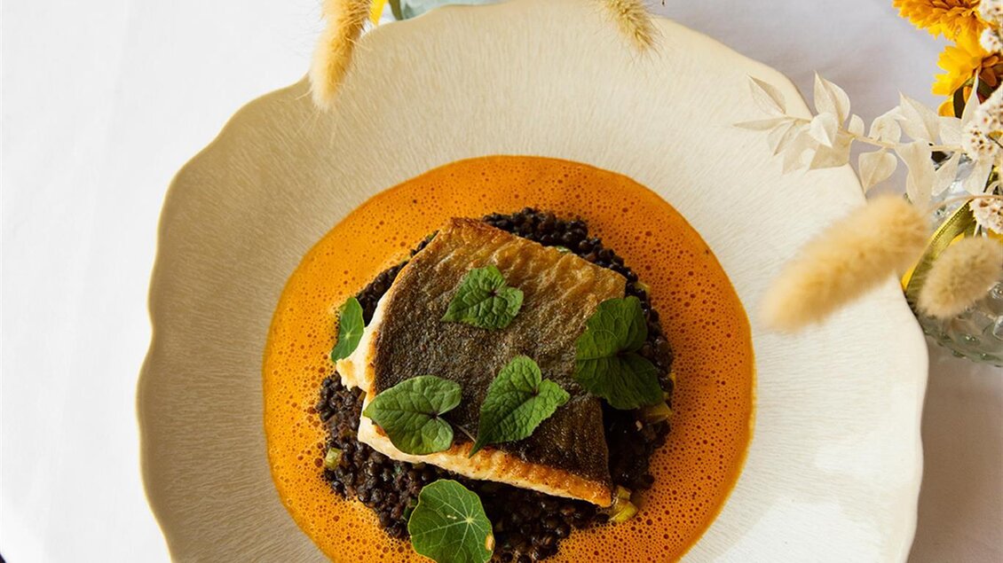 Ein kunstvoll angerichtetes Gericht mit Fischfilet auf einer Sauce und Linsen. Die Dekoration umfasst frische Kräuter und Blumen auf dem Tisch. | © Jaglhof