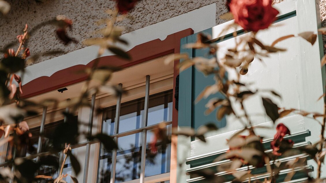 Ein Fenster mit grünen Fensterläden und Gitter ist von Rosen umgeben. Die Blüten sind rot und verleihen der Szene einen schönen Kontrast. | © Jaglhof