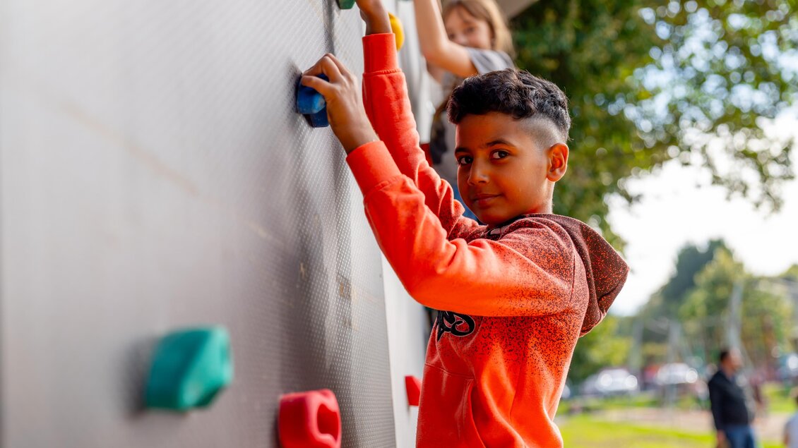 Ein Junge klettert an einer Kletterwand und hält sich an bunten Griffen fest. Im Hintergrund sind weitere Kinder und grüne Bäume zu sehen. | © © Stadtgemeinde Feldbach