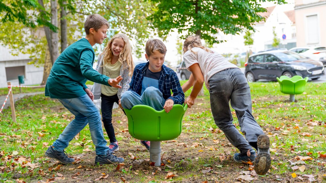Vier Kinder spielen im Freien auf einem Spielplatz. Sie drehen sich fröhlich in einem grünen Schaukelsitz. | © © Stadtgemeinde Feldbach