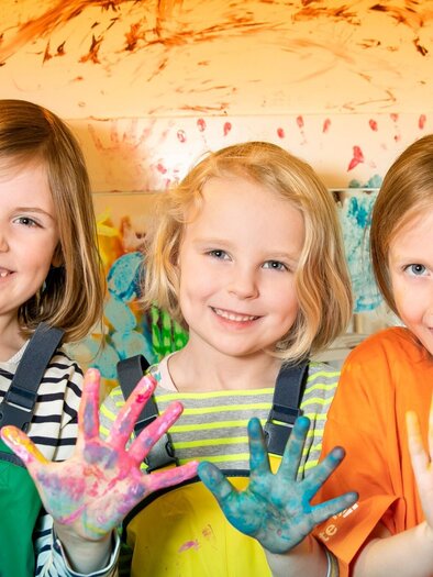 Drei lachende Kinder mit bunter Farbe an den Händen im Mitmachbereich des Kindermuseums Frida & Fred | © FRida und freD-Hannes Loske