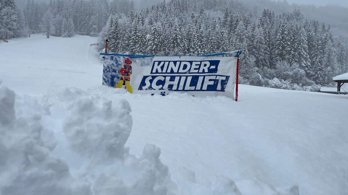 Ein verschneiter Hang mit einem Banner für den Kinderschlepplift. Im Hintergrund sind verschneite Bäume und ein grauer Himmel zu sehen. | © Kinderschilift Pölstal