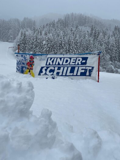 A snowy slope with a banner for the children's ski lift. In the background, there are snow-covered trees and a gray sky. | © Kinderschilift Pölstal