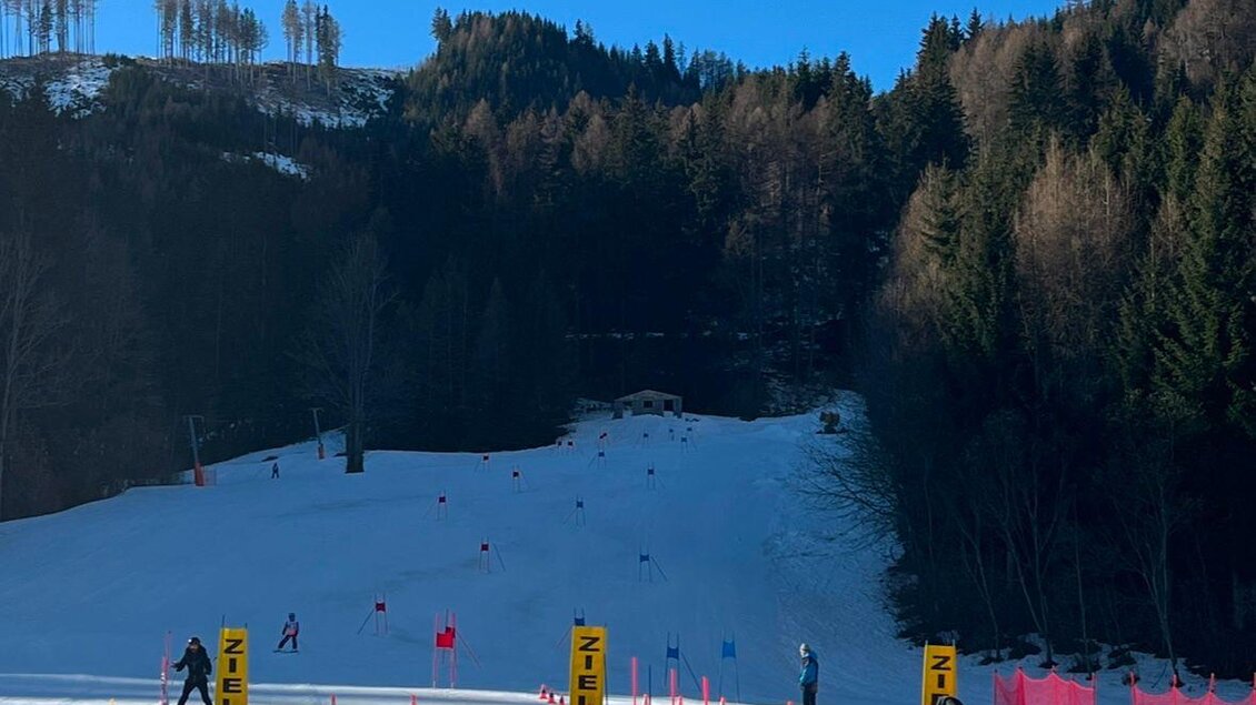 Eine schneebedeckte Skipiste mit Skifahrern in der Ferne. Der Himmel ist klar und blau, umgeben von bewaldeten Hügeln. | © Kinderschilift Pölstal