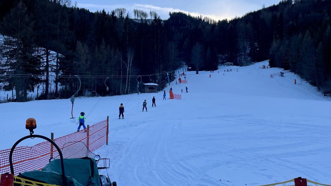 Eine verschneite Piste mit Skifahrern und einem Schlepplift im Hintergrund. Der Himmel ist klar und es sind Berge und Bäume zu sehen. | © Kinderschilift Pölstal