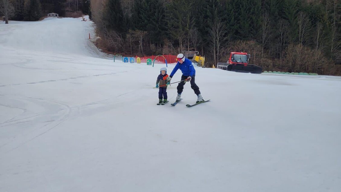 Ein Skifahrer begleitet ein Kind auf einer schneebedeckten Piste. Im Hintergrund sind Bäume und einige bunte Übungsbänder zu sehen. | © Kinderschilift Pölstal