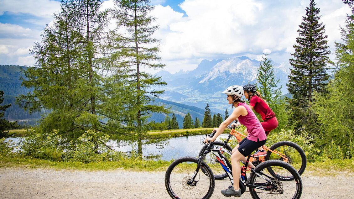 Zwei Radfahrerinnen fahren auf einem Schotterweg durch eine malerische Landschaft. Im Hintergrund sind Bäume und Berge zu sehen, unter einem klaren Himmel. | © Hauser Kaibling