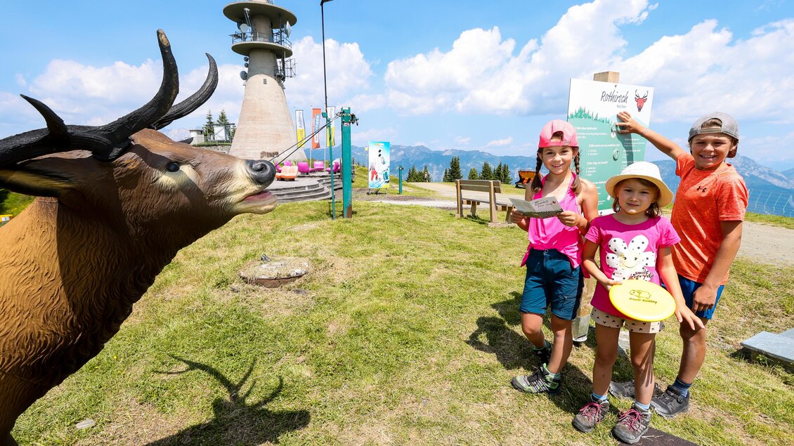 Drei Kinder posieren fröhlich vor einer Skulptur eines Ziegens. Im Hintergrund sind Berge und ein Aussichtsturm zu sehen. | © Hauser Kaibling