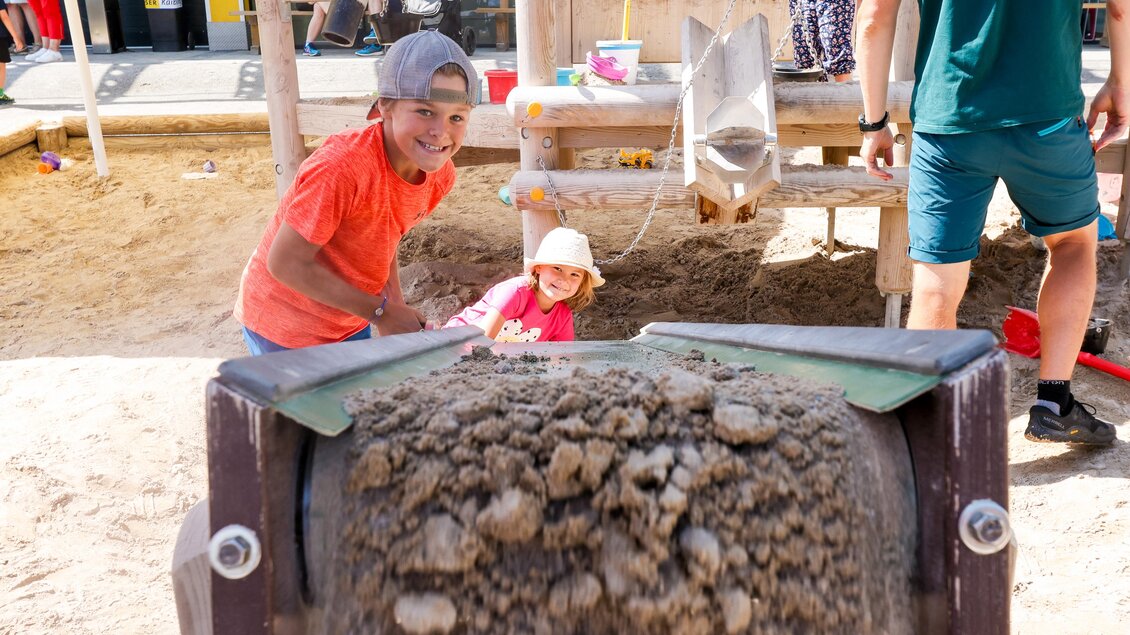 Zwei Kinder spielen im Sand. Ein Junge arbeitet mit einem Sandspielzeug, während ein Mädchen zuschaut. | © Hauser Kaibling