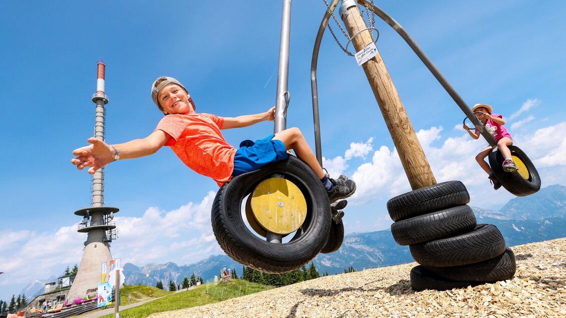 Ein Junge schwingt fröhlich auf einer Reifenschaukel mit Bergblick. Im Hintergrund ist ein Sendeturm zu sehen. | © Hauser Kaibling