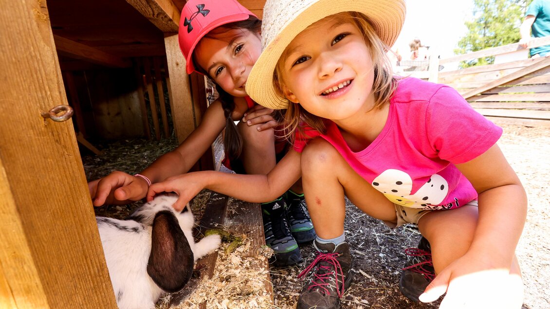 Zwei Mädchen spielen mit einem Kaninchen in einem Tiergehege. Sie lächeln und genießen die Zeit draußen. | © Hauser Kaibling
