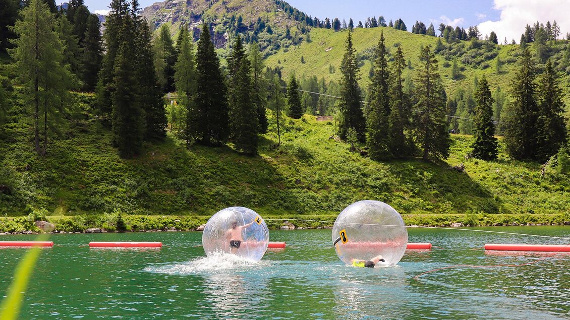 Zwei Personen schwimmen in transparenten Wasserbällen auf einem klaren grünen See. Im Hintergrund sind üppige Bäume und sanfte Hügel zu sehen. | © Hauser Kaibling